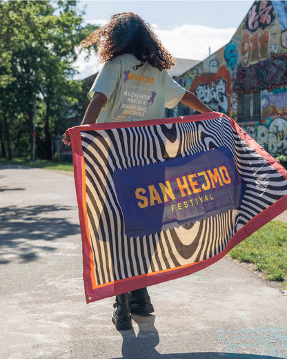 Eine Frau läuft an einer mit Graffiti besprühten Gebäudewand vorbei, Sie hält die San Hejmo Flagge ausgebreitet Die Flagge hat einen roten Rahmen und ein durchgehendes Zebra Muster. Mittig ist in lila die Form eines Tickets zu sehen. In diesem steht in Gelb "San Hejmo Festival"