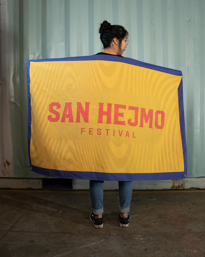 Eine Person hält eine Flagge hinter ihrem Rücken ausgebreitet in Richtung Kamera. Die Flagge hat einen lila Rahmen und einen gelben Hintergrund. Mittig stehen in Großbuchstaben die Worte "San Hejmo Festival"