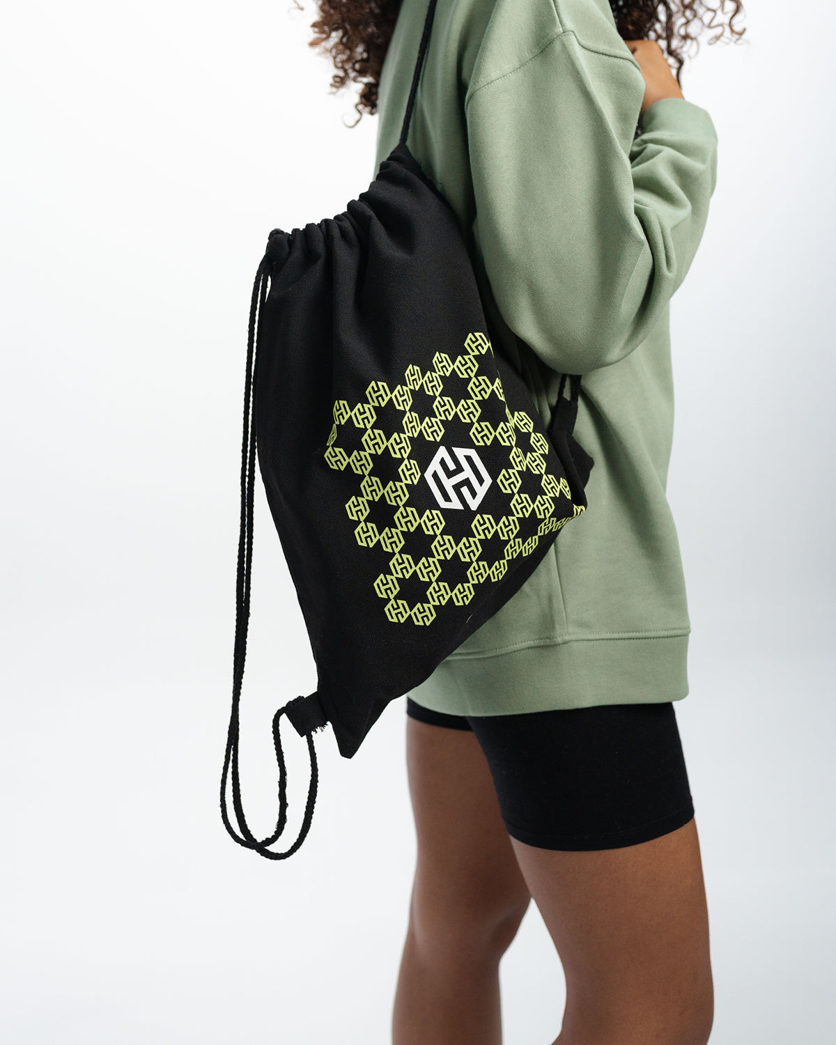 Schwarzer sportlicher Beutel mit dem in Bienenwaben angeordneten San Hejmo Logo in Mint. Mittig ist das San Hejmo in Weiß abgebildet. Eine Frau trägt ihn auf einer Schulter und steht zur Kamera gedreht.