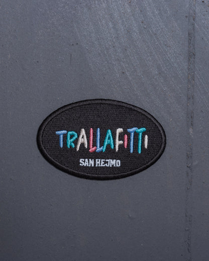 Ein Magnet der optisch aussieht wie ein Patch liegt auf einer grauen Metallwand. In jeweils einzeln gestickten bunten Farben steht dort in Großbuchstaben "Trallafitti". Die Schriftgröße ist innerhalb der Buchstaben unterschiedlich. Unter "Trallafitti" steht ebenfalls in Großbuchstaben "San Hejmo" in weiß gestickt.