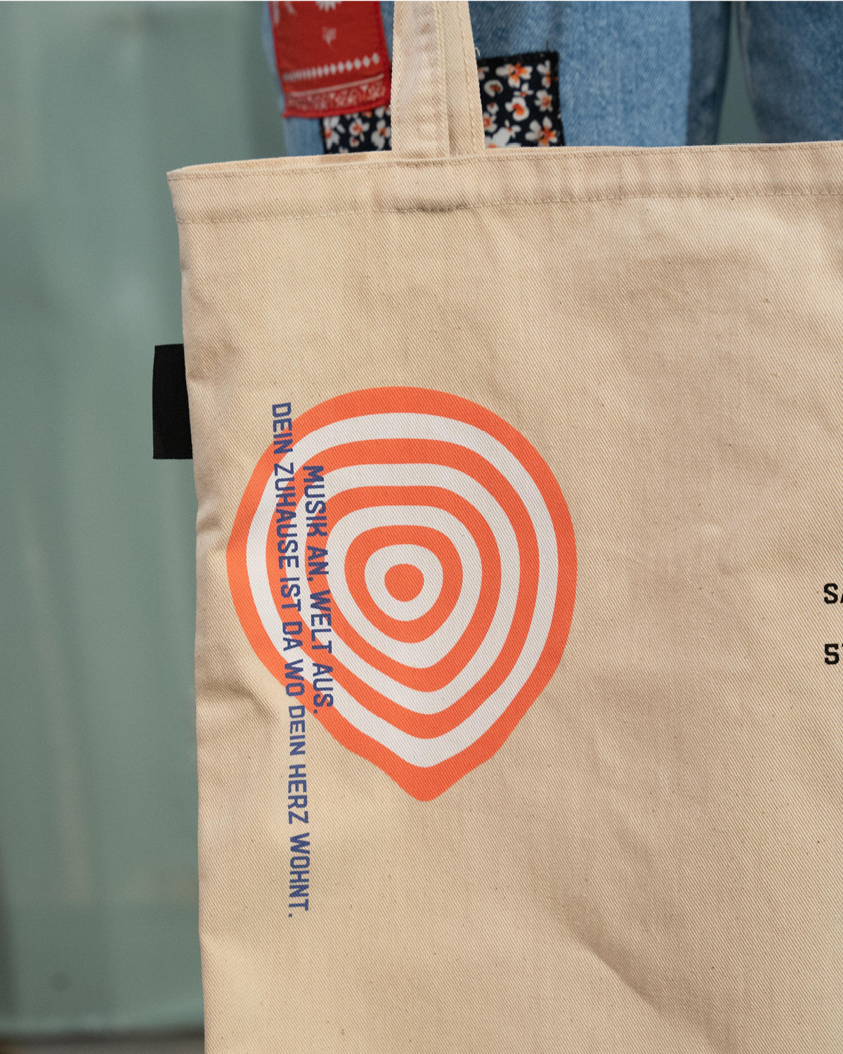 Die Rückseite des naturbelassenden Beutels mit Zoom auf die obere linke Ecke. Zu sehen ist ein Zielscheiben-artiger Kreis in weiß und orange. Am linken Rand stehen senkrecht die Worte "Musik an, Welt aus. Dein Zuhause ist da wo dein Herz wohnt".