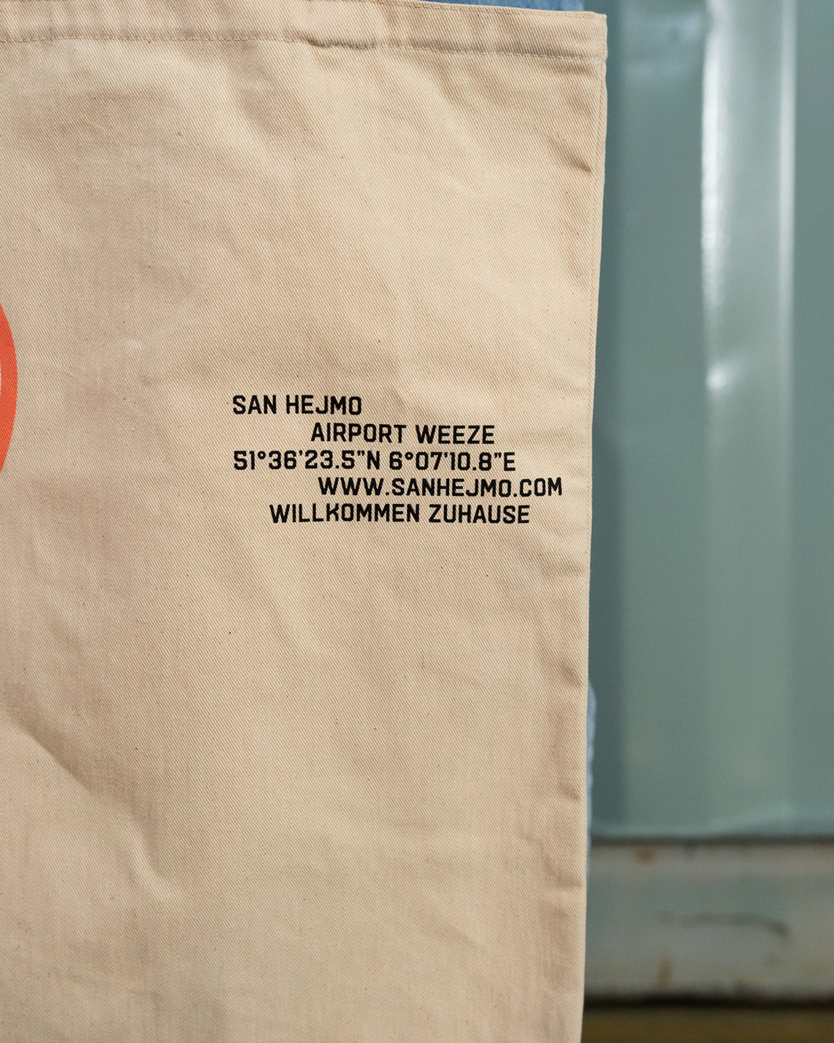 Die rechte Ecke des naturbelassenen Beutels. Zu sehen ist eine schwarze Schrift die die Geodaten von San Hejmo darstellt. Darunter ist die Internetseite und die Worte "Willkommen Zuhause" abgedruckt. 