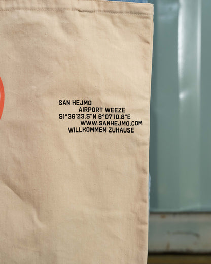 Die rechte Ecke des naturbelassenen Beutels. Zu sehen ist eine schwarze Schrift die die Geodaten von San Hejmo darstellt. Darunter ist die Internetseite und die Worte "Willkommen Zuhause" abgedruckt. 