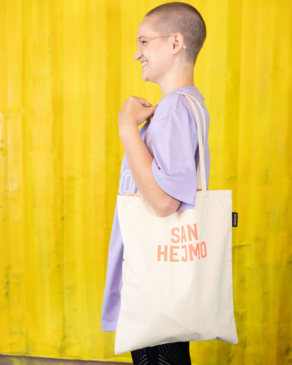 Eine Person trägt einen naturbelassenen, beigen Beutel und ist seitlich gedreht, 
damit der Beutel gerade zur Kamera fällt. Mittig auf dem Beutel ist der Schriftzug "San Hejmo" in orange mit kleinem weißen Rahmen um jeden Buchstaben gedruckt. Die Person ist vollständig im Bild zu sehen.
