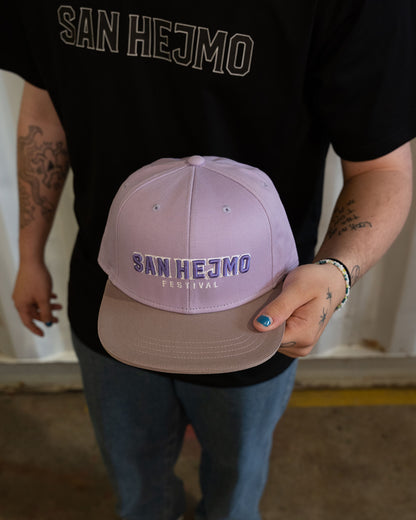 Eine Person trägt die lilac San Hejmo Basecap in der Hand, so dass die Cap aus der  Vogelperspektive zu sehen ist. Ein großer "San Hejmo" Stick in Lila mit weißen Rahmen um die einzelnen Buchstaben, sitzt vorne mittig. 