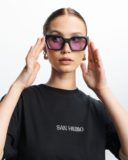 Eine Sonnenbrille mit einem eckig gehaltenen schwarzem Rahmen und rosa Gläsern. Sie wird von einer Frau getragen. 