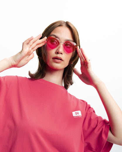 Eine Frau trägt ein pinkes Oberteil und die herzförmige rosa Sonnenbrille von San Hejmo. Sie hält diese rechts und links mit den Fingerspitzen fest.