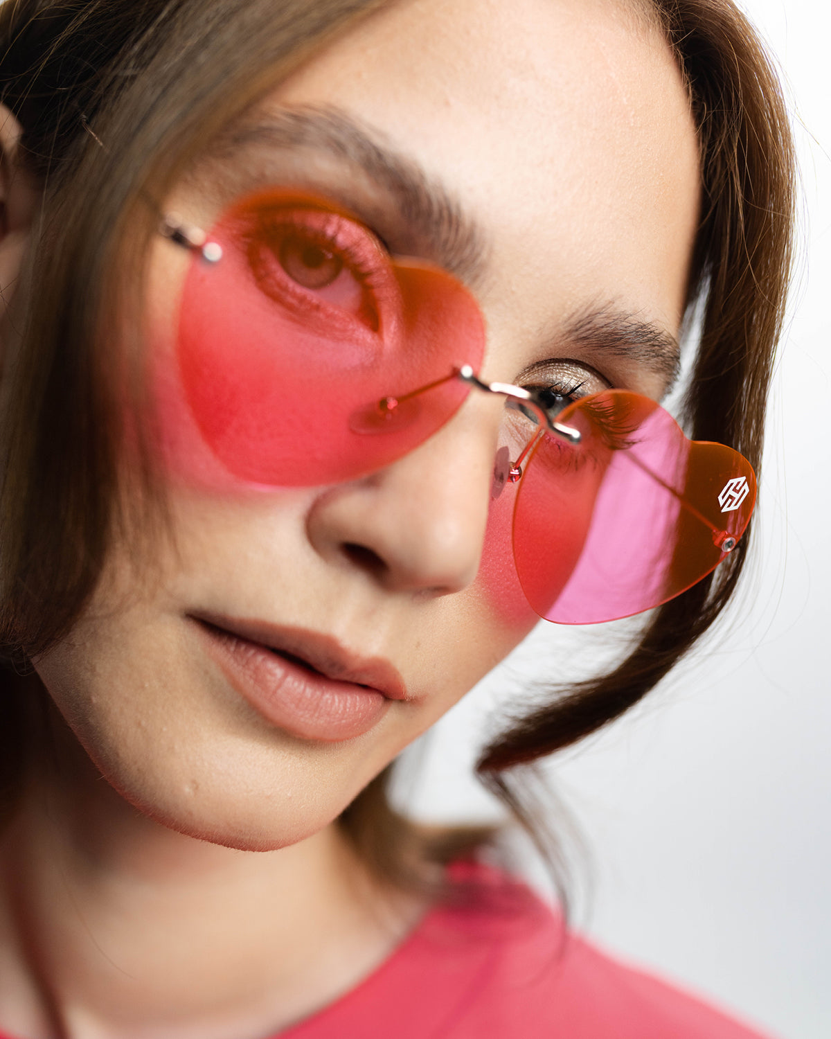 Eine Nahaufnahme einer Frau, die die San Hejmo Sonnenbrille in Herzform trägt. Die Gläser sind rosa, die Brücke und die Bügel sind in Gold. Am oberen rechten Glasrahmen ist das San Hejmo Logo in weiß gedruckt. 