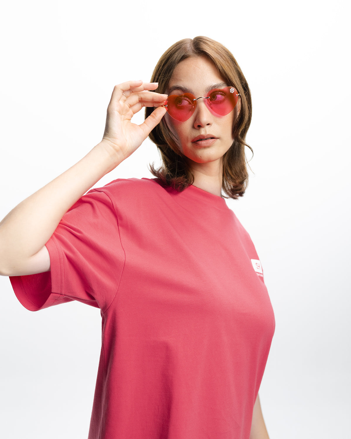 Eine Frau mit pinkem Oberteil steht mit gedrehtem Oberkörper zur Kamera. Sie hält mit Ihrer linken Hand die Herzförmige rosa Sonnenbrille von San Hejmo fest. 