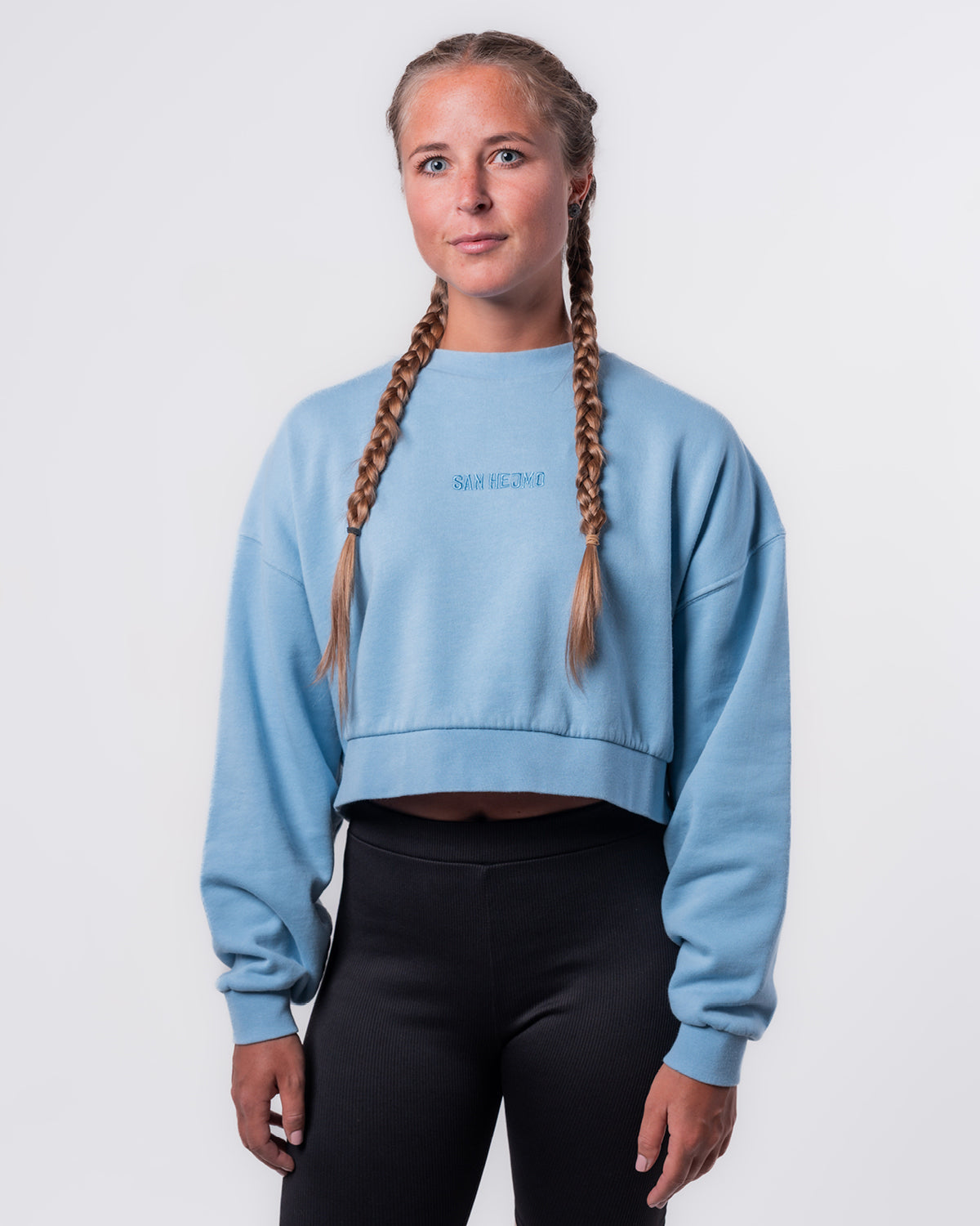 Zu sehen ist eine junge Frau, sie trägt eine schwarze Hose und den hellblauen Cut Out Sweater und steht vor einer weißen Wand. Ihre Haare sind in zwei Zöpfe geflochten. Der Sweater ist oversized geschnitten und endet in der Länge auf Hüfthöhe. In der Mitte des Sweaters ist der Schriftzug "San Hejmo" in hellblau gestickt.