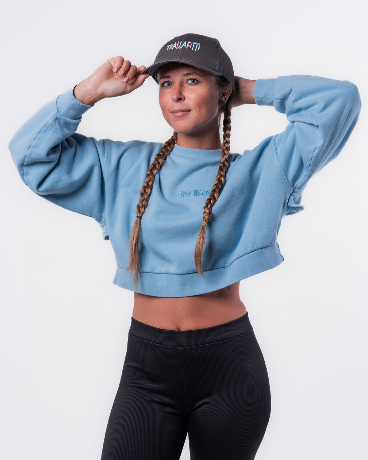 Eine junge Dame mit geflochtenen langen Haaren vor weißem Hintergrund. Sie trägt eine schwarze Hose, den hellblauen Cut Out Sweater und eine Anthrazitfarbene Cap mit der bunten Aufschrift "Trallafitti".