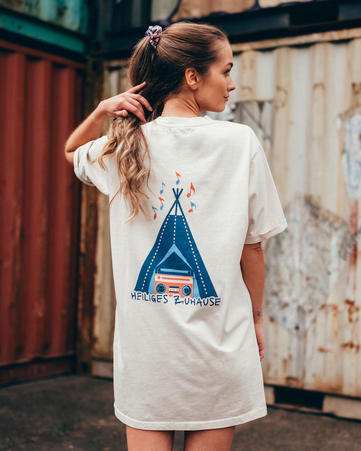 Eine junge Frau steht vor einer Containerwand mit dem Rücken zur Kamera. Sie trägt ein weißes T-Shirt Kleid mit einem großen Aufdruck auf dem Rücken. Dieser bildet ein offenes blaues Tipi-Zelt mit einer Coral farbigen Musikbox davor ab. über dem Tipi fliegen Musiknoten in blau und orange. unter dem Tipi steht in Großbuchstaben "Heiliges Zuhause".