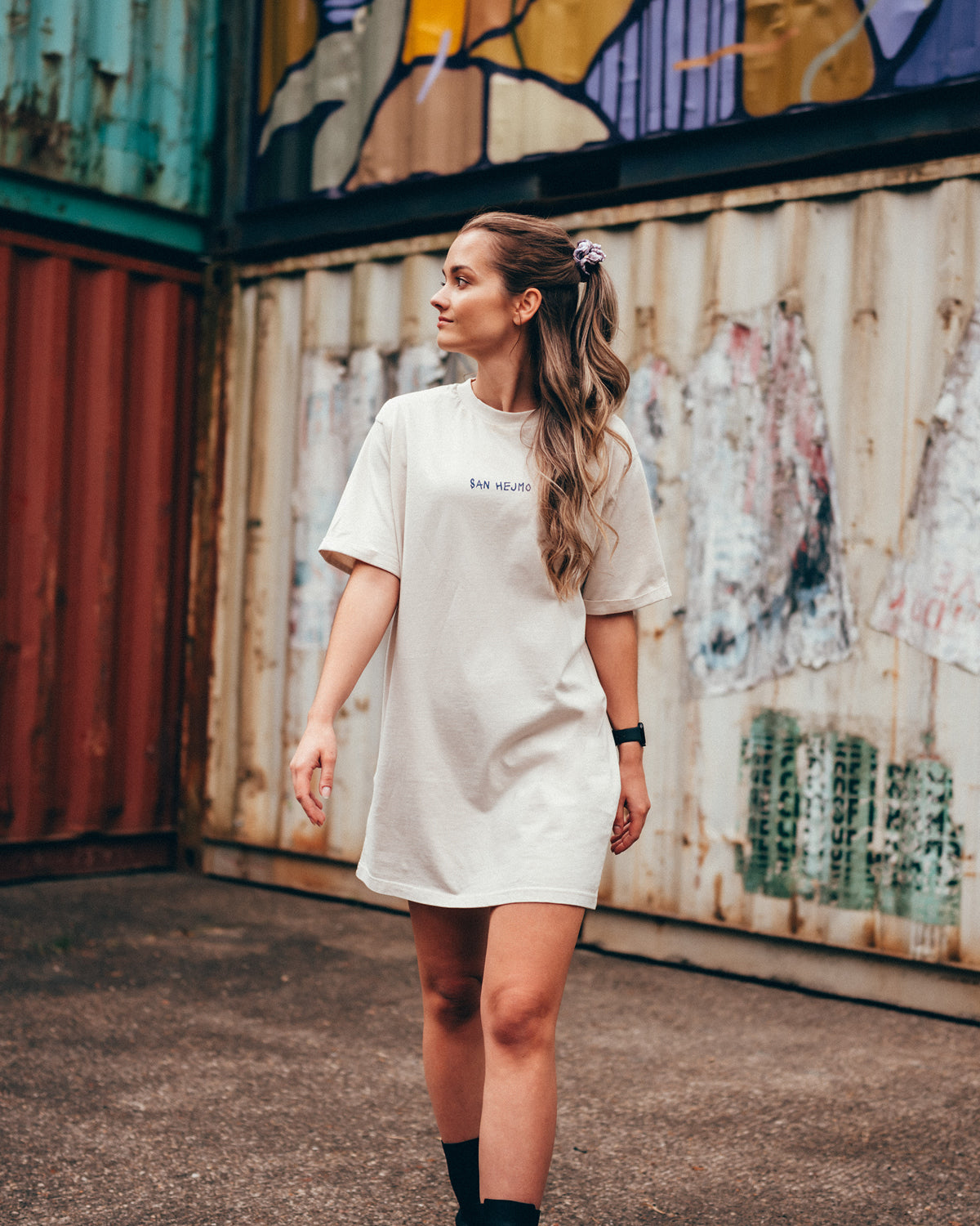 Eine junge Frau steht vor einer Containerwand mit dem Rücken zur Kamera. Sie trägt ein weißes T-Shirt Kleid mit einer einfachen Schrift vorne auf Brusthöhe. Darauf steht in Großbuchstaben "San Hejmo" in dunkler Schrift. 