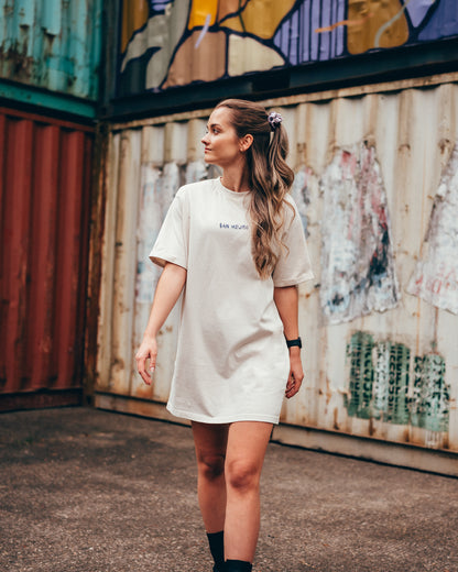 Eine junge Frau steht vor einer Containerwand mit dem Rücken zur Kamera. Sie trägt ein weißes T-Shirt Kleid mit einer einfachen Schrift vorne auf Brusthöhe. Darauf steht in Großbuchstaben "San Hejmo" in dunkler Schrift. 