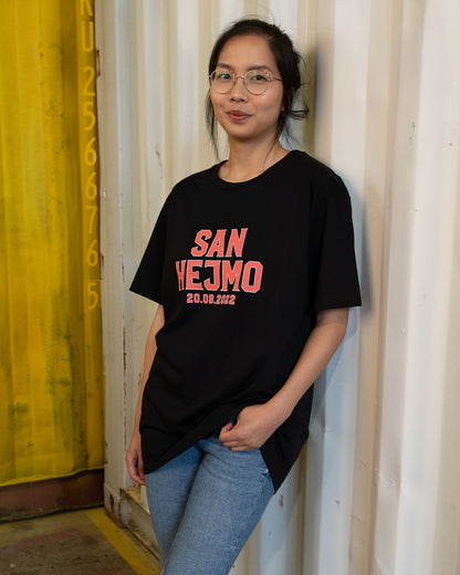 Das schwarze Line-up T-Shirt 2022, getragen von einer Frau. Es trägt die orangene Aufschrift "San Hejmo 20.08.2022". Die Schrift hat einen weißen Rahmen
