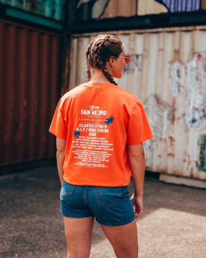 Eine junge Frau steht vor einer Containerwand, mit dem Rücken zur Kamera. Sie trägt eine kurze Jeanshose und ein Coral farbenes T-Shirt. Auf dem T-Shirt ist das Line-up des Jahres 2025 zu sehen. Oben rechts und mittig links ist die Schrift mit jeweils einem blauen Farbklecks hinterlegt. 