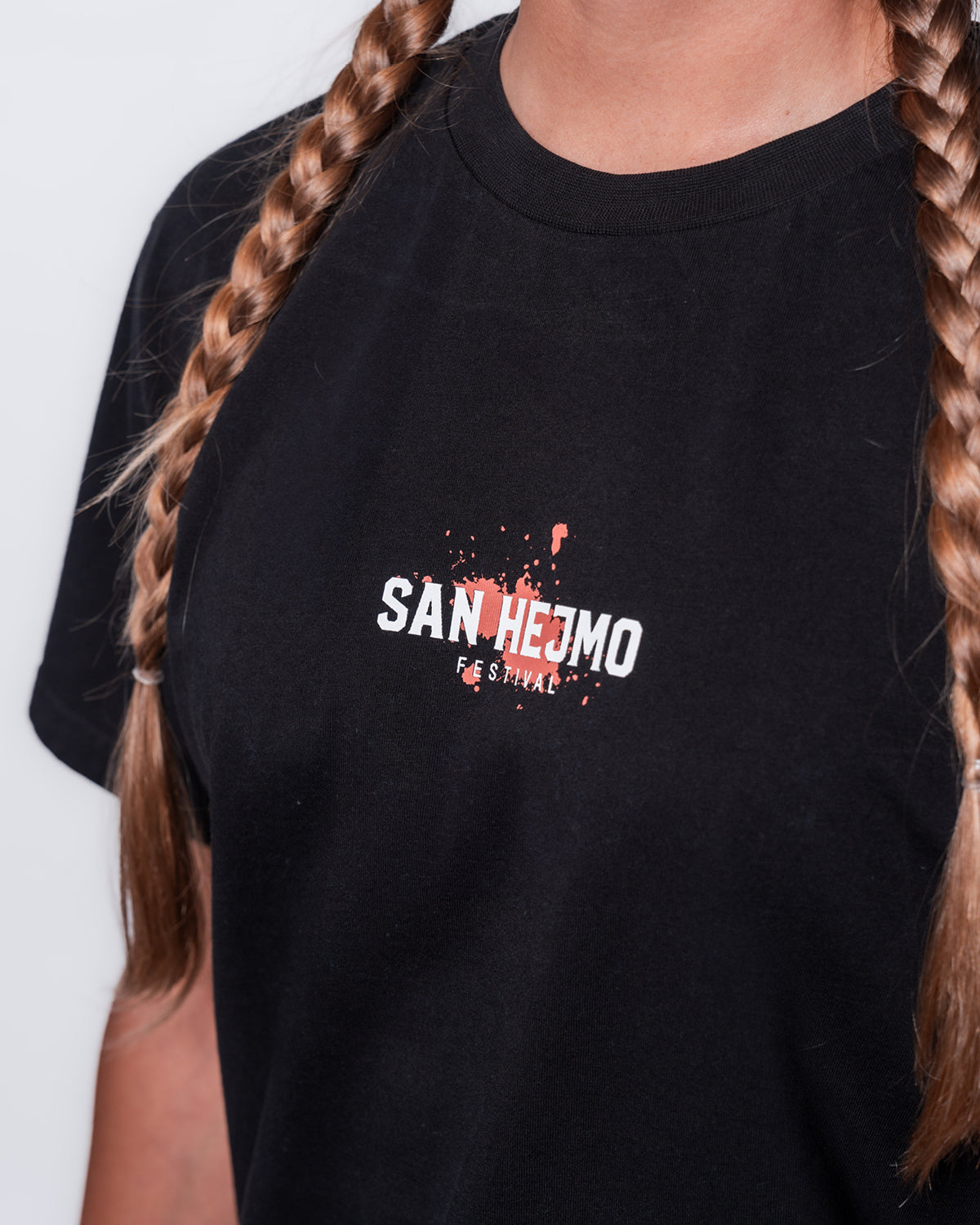 Eine Nahaufnahme des Brustprints des San Hejmo Line-up 2025 Oberteils. In weißer Schrift und Großbuchstaben steht "San Hejmo" und eine Zeile darunter in kleinerer Schrift "Festival". Der Print ist mit einem Coral farbigen Klecks hinterlegt.