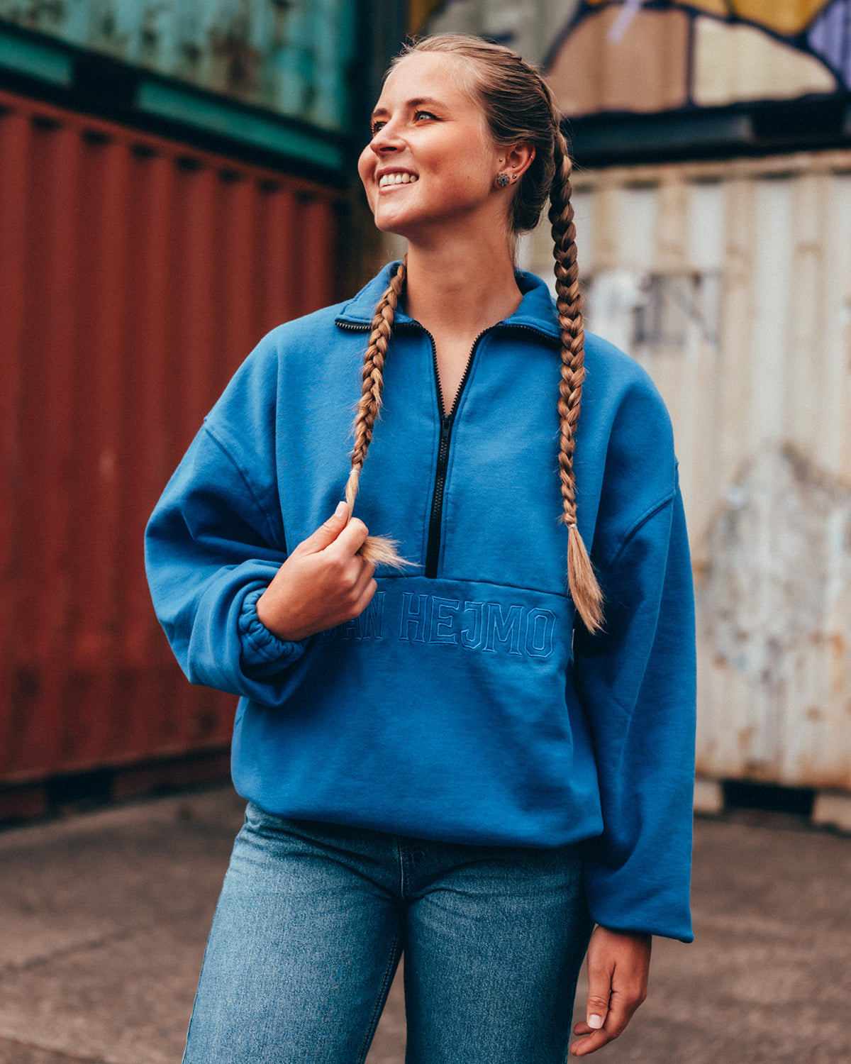 Eine junge Frau steht vor einer Containerwand und trägt eine blaue Jeans, den blauen San Hejmo Troyer Sweater und hat Ihre Haare in zwei Zöpfen geflochten. Der Troyer hat einen oversized Schnitt. Mittig längs zieht sich eine Naht über den Oberkörper, unter Ihr sind in Großbuchstaben die Worte San Hejmo gestickt. Mittig von dieser Naht ist ein schwarzer Half-Zip eingenäht. Der vorhandene Kragen ist umgeklappt.