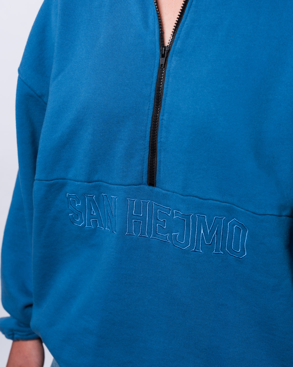 EIne Nahaufnahem des Half-Zip und des hellblauen Sticks. Unter der mittigen Längsnaht ist in Großbuchstaben "San Hejmo" gestickt. Der schwarze Half-Zip ist mittig bis zur Längsnaht und geht bis zum Ende des Kragens. Der Kragen des Troyers ist umgeklappt. 