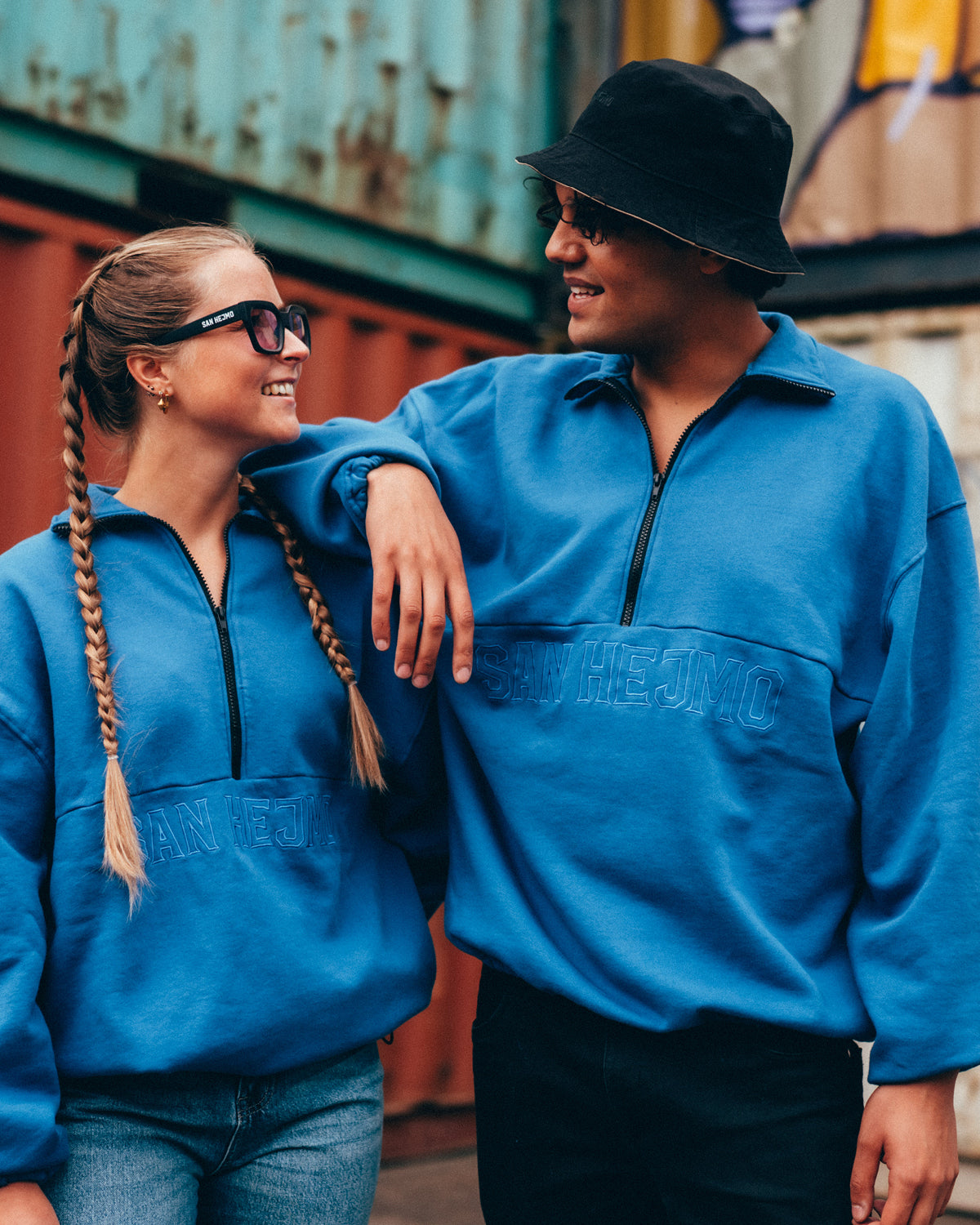 Eine Frau und ein Mann tragen beide den blauen San Hejmo Troyer und stehen in einer lässigen Pose. Unter der mittigen Längsnaht ist in Großbuchstaben "San Hejmo" gestickt. Der schwarze Half-Zip ist mittig bis zur Längsnaht und geht bis zum Ende des Kragens. Der Kragen des Troyers ist bei beiden umgeklappt. 